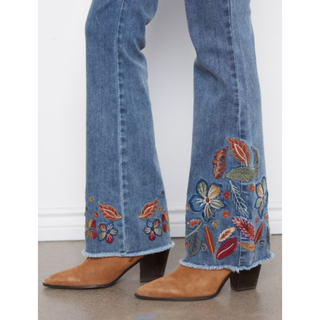 Close-up of a person's legs wearing blue denim flare jeans with colorful floral embroidery near the hem and tan suede heeled boots.