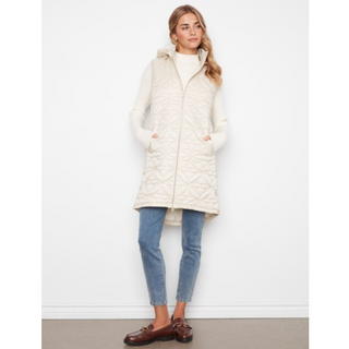 A woman modeling a long cream quilted puffer vest with a hood and front zipper, worn over a cream turtleneck, blue jeans, and brown loafers against a plain white wall.