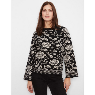 A woman modeling a black long-sleeve crewneck sweater with an intricate black and white floral and vine pattern, paired with black pants, against a plain light gray background.