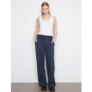 A woman stands against a plain white wall wearing a white sleeveless top and navy wide-leg pants, with her hands in the pockets of the pants.