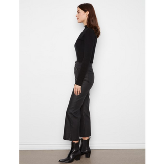 A woman stands in profile wearing a fitted black ribbed long-sleeve top, black wide-leg cropped pants, and black ankle boots against a plain white wall.