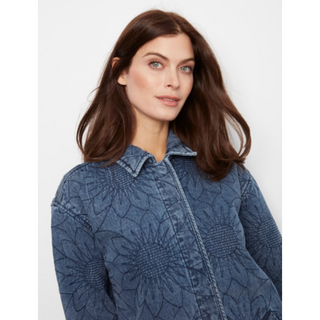 A woman with long brown hair wearing a textured blue denim jacket with a subtle sunflower pattern, photographed against a light gray background.