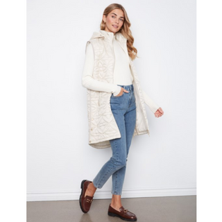 A woman models a long cream quilted sleeveless puffer vest over a white ribbed knit top, paired with blue jeans and brown loafers, standing against a plain white wall.
