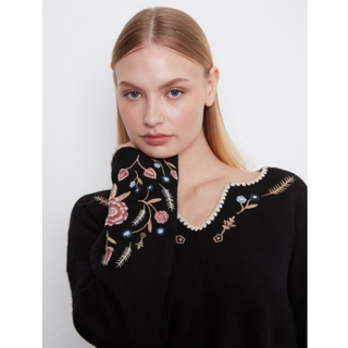 A woman wearing a black knit sweater with detailed floral embroidery around the neckline and sleeve, posed against a light gray studio background.