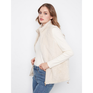 A woman wearing a cream-colored quilted sleeveless vest over a matching cream long-sleeve top and blue jeans, photographed against a plain light gray studio background.