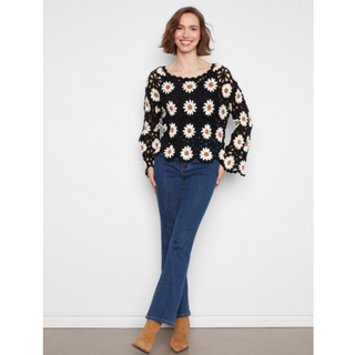 A woman modeling a black long-sleeve top with a white and yellow daisy pattern, paired with blue jeans and tan ankle boots, standing against a white wall.