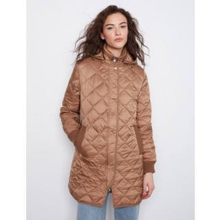 A woman wearing a long quilted hooded puffer coat in a warm camel brown color, paired with light blue jeans, photographed against a plain light gray studio background.