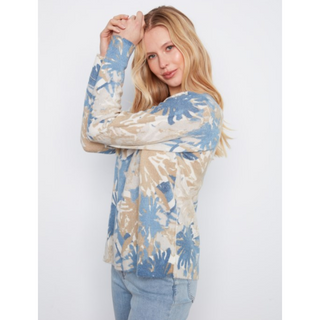 A woman modeling a long-sleeve knit top with a blue and beige floral pattern, paired with light-wash blue jeans, against a plain light gray background.