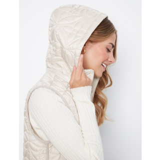 A woman in profile wearing a cream-colored quilted puffer vest over a matching ribbed knit sweater, set against a plain light gray studio background.