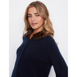 A woman wearing a navy blue knit top with a brown faux-fur collar, photographed against a plain light gray studio background.