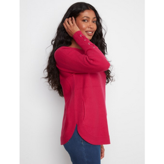 A woman modeling a bright pink long-sleeve knit top with lace-up detail on the cuffs, paired with blue jeans, against a plain light gray background.