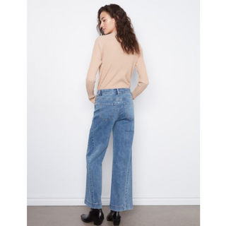 A woman stands with her back to the camera wearing a beige ribbed turtleneck sweater, wide-leg blue denim jeans, and black ankle boots against a plain white wall.
