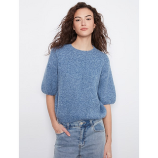 A woman wearing a heathered blue short-sleeve knit sweater and light-wash blue jeans, standing against a plain light gray studio background.