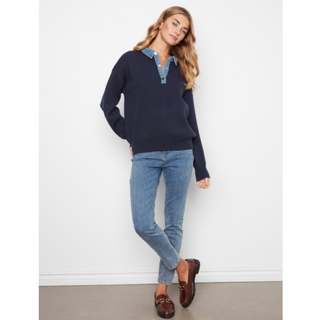 A woman modeling a navy blue long-sleeve knit sweater with a light blue denim polo-style collar, paired with light blue jeans and brown loafers, standing against a white wall.