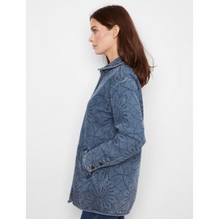 A woman shown in side profile wearing a blue denim jacket with an allover tonal leaf or vine pattern, set against a plain light gray studio background.