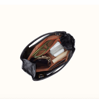 An overhead view of an open black handbag showing its interior organization with various accessories including a brown organizer insert, a pair of green sunglasses, a black wallet, a white cord, and a beige stick, set against a plain off-white background.