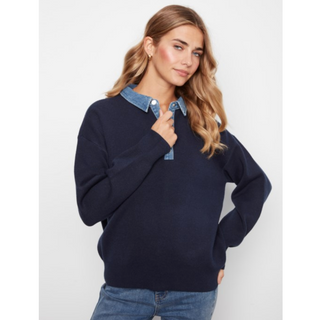 A woman modeling a navy blue knit sweater with a light blue denim collar, paired with blue jeans, against a plain light gray background.