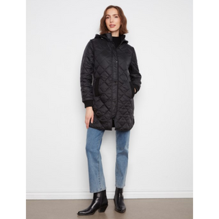 A woman modeling a black quilted hooded jacket, paired with blue jeans and black ankle boots, standing against a plain white wall.