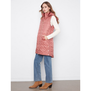 A woman modeling a dusty rose pink quilted sleeveless hooded puffer vest over a white long-sleeve top, paired with blue jeans and tan ankle boots, standing against a plain white wall.