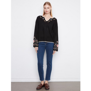 A woman modeling a black long-sleeve blouse with floral embroidery on the sleeves and neckline, paired with blue jeans and brown loafers, standing against a white wall.