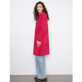A woman modeling a bright pinkish-red long knit jacket, paired with light-wash wide-leg jeans and brown loafers, standing against a plain white wall.
