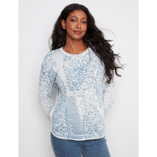 A woman modeling a light blue and white patterned knit sweater with long sleeves, paired with blue jeans, against a plain light gray background.