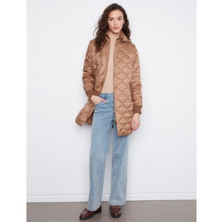 A woman stands against a plain white wall wearing a tan quilted puffer jacket over a beige top, light blue wide-leg jeans, and dark brown loafers.