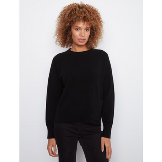 A woman with curly light brown hair wearing a black long-sleeve knit sweater and black pants, standing against a light gray studio background.