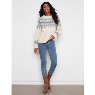 A woman modeling a cream knit sweater with a gray and blue Nordic pattern, paired with blue skinny jeans and brown boots, standing against a white wall.
