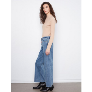 A woman modeling a beige ribbed long-sleeve top and wide-leg blue jeans with black ankle boots, standing against a plain white wall.