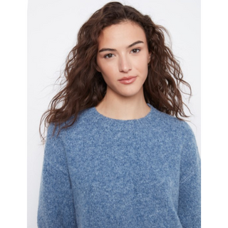 A woman with wavy brown hair wearing a heathered blue knit crewneck sweater, shown from the chest up against a plain light gray background.
