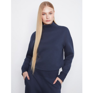 A blonde model wearing a navy blue ribbed turtleneck sweater and matching navy knit bottoms, standing against a light gray studio background.