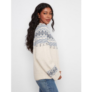 A woman wearing a cream knit sweater with blue and gray geometric Fair Isle patterns on the shoulders, sleeves, and hem, paired with blue jeans, photographed against a light gray studio background.