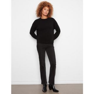A model with curly light brown hair stands against a white wall wearing a black crewneck sweater, black pants, and black ankle boots.