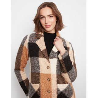 A woman wearing a fuzzy oversized plaid coat in beige, brown, black, and cream over a black turtleneck, photographed against a light gray studio background.
