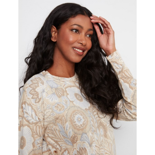 A woman with long, wavy black hair wearing a beige and white floral patterned long-sleeve top, posing against a light gray background.