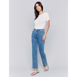 A woman modeling a white short-sleeve top and light blue straight-leg jeans with white strappy sandals, standing against a light gray studio background.