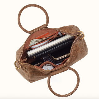 An open brown embossed leather handbag with gold-tone hardware, shown from above on a white background. The bag is filled with wallets, cash, a notebook, and keys, and features two circular top handles.