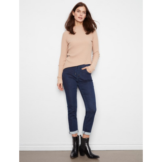 A woman modeling a beige ribbed turtleneck sweater, dark blue cropped jeans with rolled cuffs, and black ankle boots against a plain white wall.