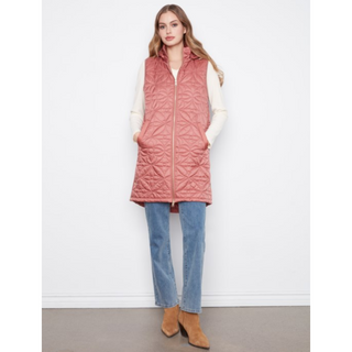 A woman stands against a plain white wall wearing a sleeveless quilted puffer vest in a dusty rose pink color, layered over a cream long-sleeve top, blue jeans, and tan ankle boots.