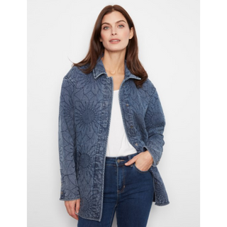 A woman modeling a blue denim jacket with a tonal sunburst floral pattern, worn open over a white top and dark blue jeans, against a light gray studio background.