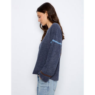 A woman wearing a heathered navy blue knit sweater with light blue and brown stripe details on the sleeves, paired with light-wash jeans, shown from a side profile against a plain white background.