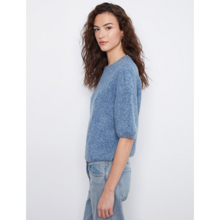 A woman with long, wavy brown hair is shown from the side, wearing a short-sleeved, textured knit sweater in a heathered medium blue color, paired with light-wash jeans, against a plain light gray background.