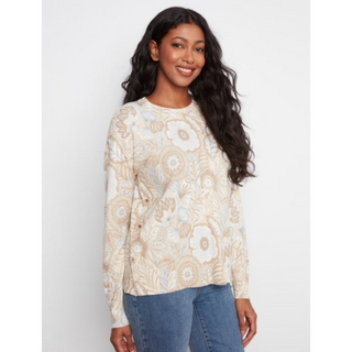 A woman modeling a cream and beige floral patterned knit sweater with blue jeans, standing against a light gray studio background.