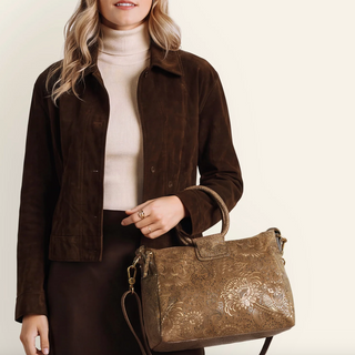 A woman wearing a brown suede jacket and cream turtleneck holds a brown embossed leather handbag with a top handle against a plain light beige background.