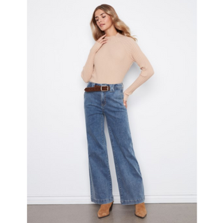 A woman modeling a beige ribbed long-sleeve mock neck top with wide-leg blue jeans, a brown belt, and tan ankle boots against a white wall.