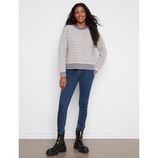 A woman modeling a striped crewneck sweater with blue jeans and black boots, standing against a plain white wall.
