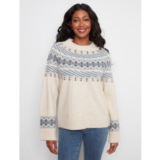 A woman wearing a cream-colored knit sweater with a blue and gray Nordic/Fair Isle pattern across the chest and cuffs, paired with blue jeans, against a light gray background.