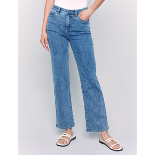 A model wearing a pair of medium-wash blue denim jeans with a straight-to-wide leg fit, paired with white slide sandals, shown against a light gray studio background.
