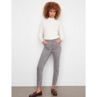 A woman with curly brown hair stands against a white wall wearing a white mock-neck long-sleeve top, gray slim-fit jeans, and brown leather loafers.